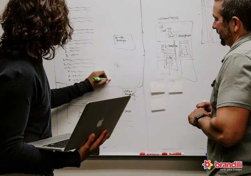 mulher segurando um notebook com uma mão, enquanto escreve no quadro para um outro homem