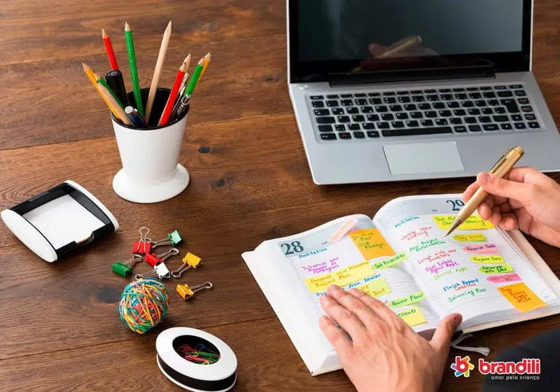 pessoa trabalhando em uma mesa com um caderno, notebook, canetas e itens de escritório em cima.