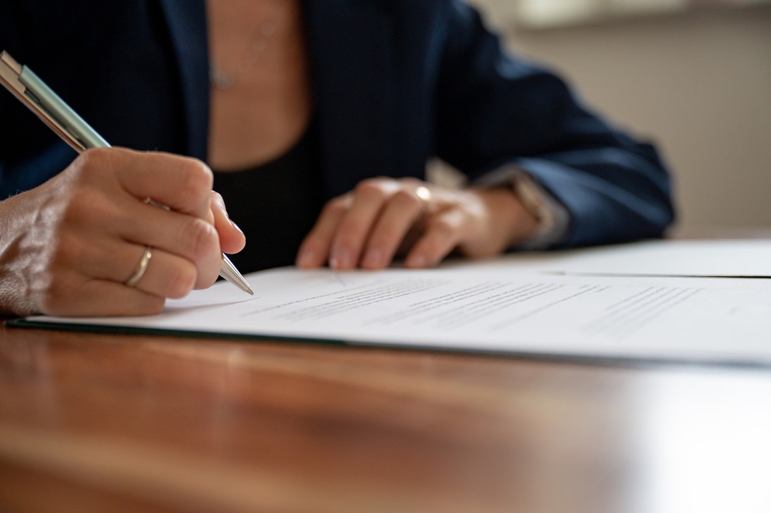 mão de uma mulher, enquadrada sem mostrar o rosto, assinando documento sob uma mesa de madeira